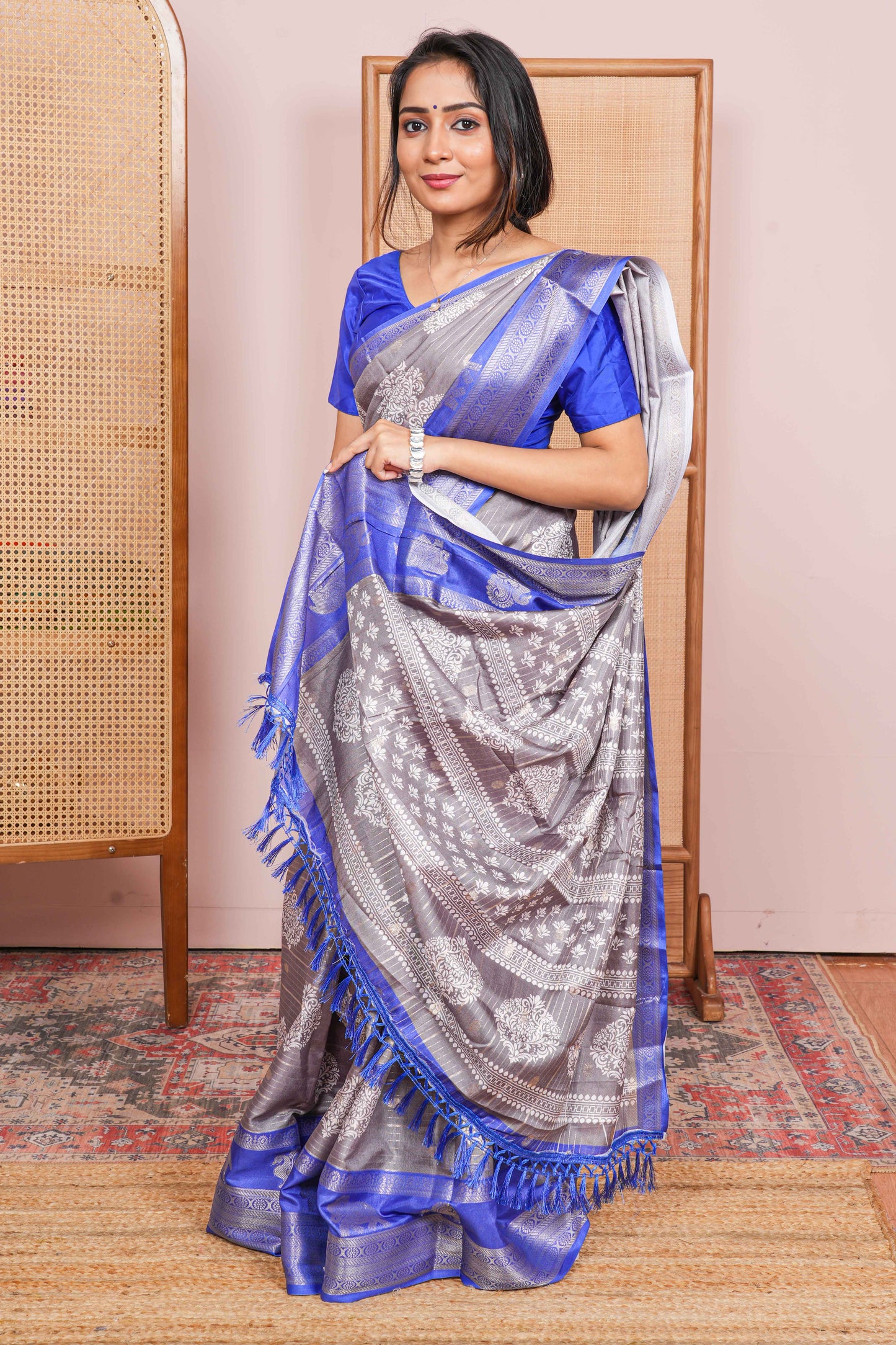 Woman wearing a blue and gray saree standing in a room with a wooden chair and pink wall.
