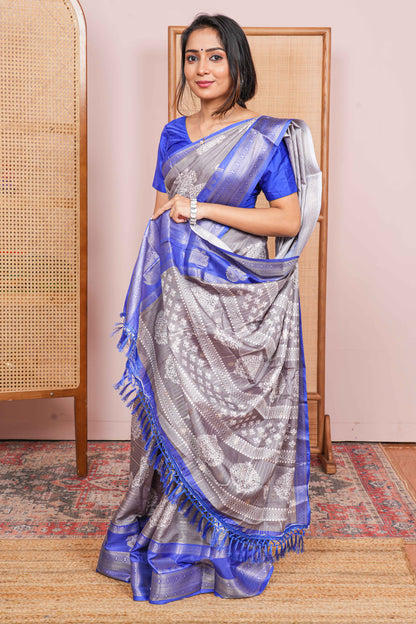 Woman wearing a blue and gray saree standing in a room with a wooden chair and pink wall.